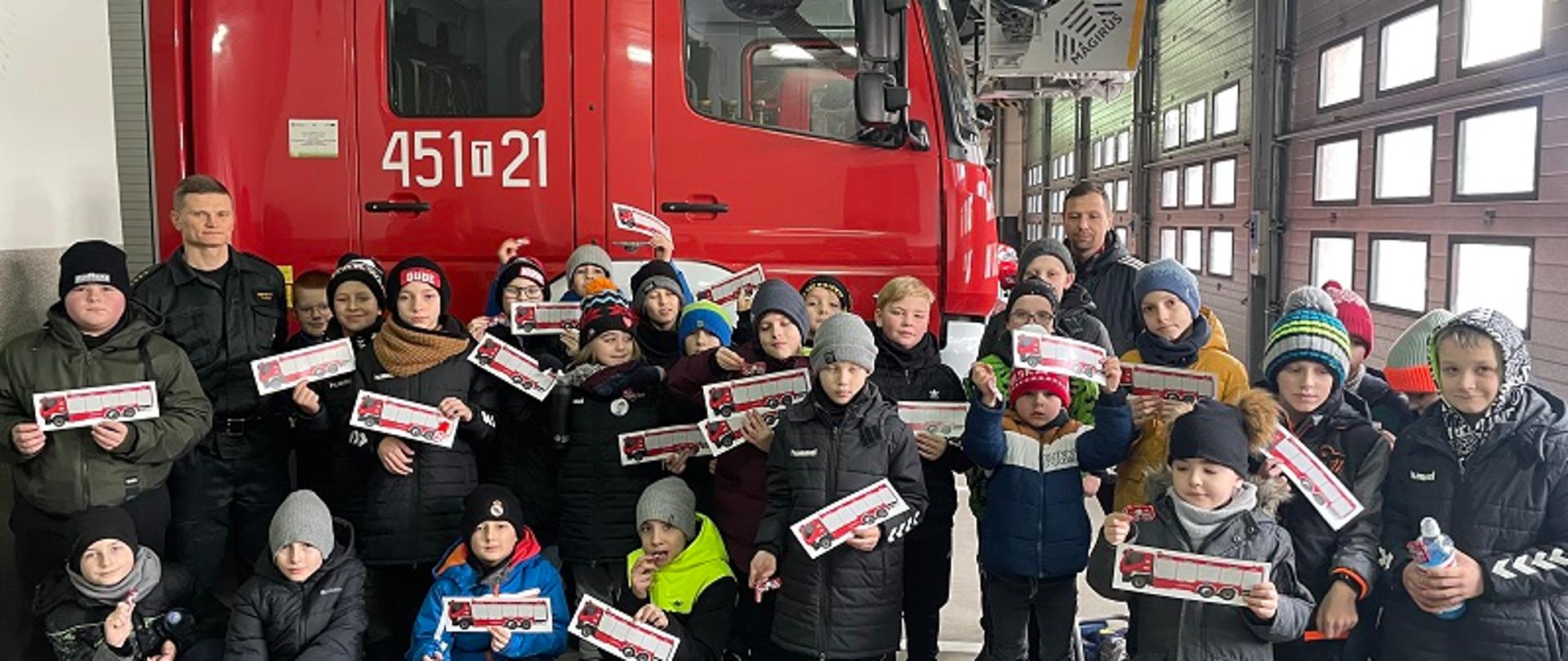 Na zdjęciu widać chłopców w wieku 5-12 lat, którzy z upominkami w rękach od strażaków pozują do zdjęcia grupowego w garażu w JRG Nr 1 w Ostrowcu Św. W tle zdjęcia samochód ratowniczo-gaśniczy