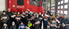 Na zdjęciu widać chłopców w wieku 5-12 lat, którzy z upominkami w rękach od strażaków pozują do zdjęcia grupowego w garażu w JRG Nr 1 w Ostrowcu Św. W tle zdjęcia samochód ratowniczo-gaśniczy