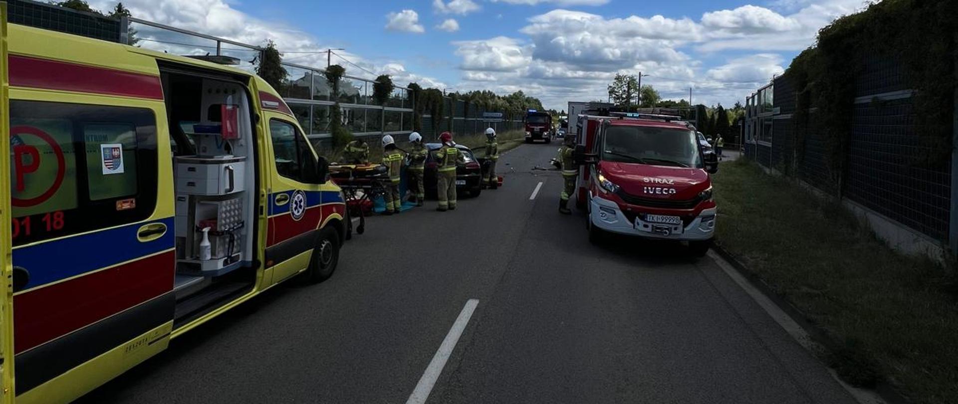 Zdjęcie przedstawia miejsce wypadku: z lewej strony ambulans, za którym trwa akcja ratownicza, a z prawej stoi samochód pożarniczy.