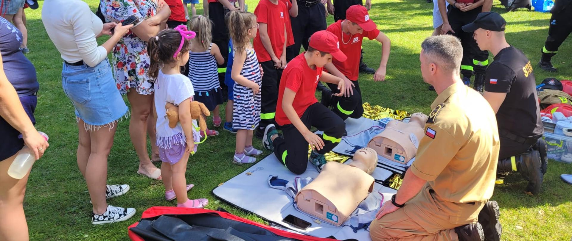 Po prawej strażacy omawiający zagadnienie, przed nimi na matach leżą fantomy do nauki resuscytacji przy których kucają uczestnicy festynu. Za nimi stojący widzowie