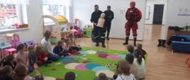 Zdjęcie wykonane jest w sali lekcyjnej i przedstawia grupę dzieci siedzących na kolorowym dywaniku, które słuchają strażaków. Dwóch strażaków ubranych jest w ubranie koszarowe, jeden z nich trzyma manekina do udzielania pierwszej pomocy. Trzeci strażak ubrany jest w ubranie wypornościowe do pracy w wodzie. W sali znajdują się zabawki dla dzieci, a przez okna w tle zdjęcia widoczny jest plac zabaw.