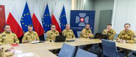 Sześciu funkcjonariuszy Państwowej Straży Pożarnej w mundurach w kolorze piaskowym siedzi przy stole konferencyjnym. Przed nimi laptopy, notatniki i dokumenty. W tle stoją flagi Polski i Unii Europejskiej oraz baner z napisem „Komenda Główna Państwowej Straży Pożarnej”.