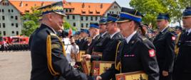 Na zdjęciu widać odznaczonych i wyróżnionych funkcjonariuszy PSP w mundurach wyjściowych. Zdjęcie wykonane w ciągu dnia. W tle widać krzewy i drzewa z zieloną koroną. Za drzewami, w centralnym punkcie budynek.