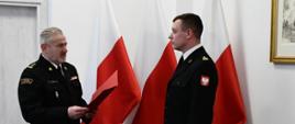 Strażak w mundurze wyjściowym stoi i trzyma teczkę przed nim stoi strażak w mundurze wyjściowym za nimi ustawione są trzy flagi Polski na ścianie wisi obraz.