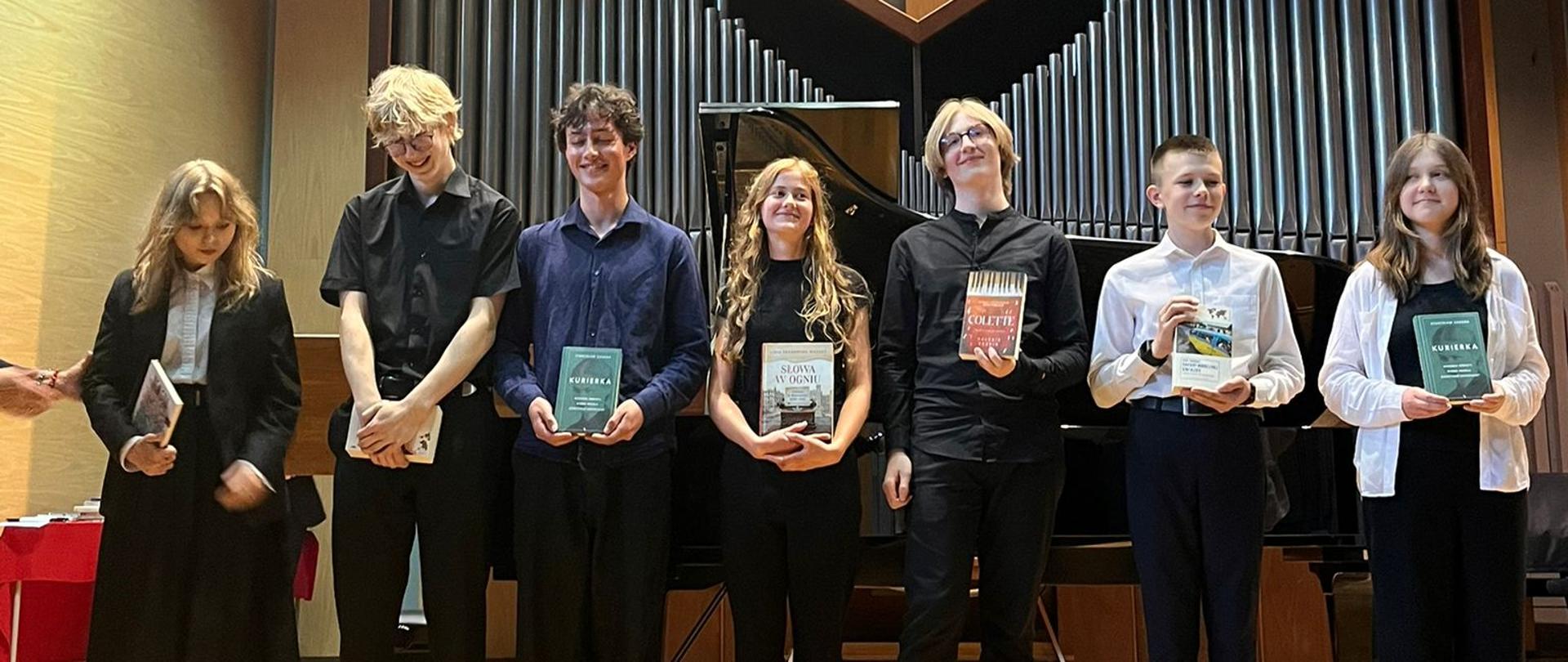 Na zdjęciu widzimy sześciu uczniów stojących na scenie w sali koncertowej. W tle znajduje się duży instrument – organy piszczałkowe oraz fortepian. Osoby ustawione są w szeregu, zwrócone twarzą do obiektywu. Każda z nich trzyma w rękach książkę, k – są to nagrody za osiągnięcia szkolne lub artystyczne. Wszyscy są elegancko ubrani – dominują stroje galowe w kolorach czerni i bieli. Oświetlenie sceniczne jest jasne, a sufit i reflektory nadają wnętrzu nowoczesny charakter. Zdjęcie emanuje atmosferą uroczystości i dumy, prawdopodobnie wykonano je podczas zakończenia roku szkolnego w szkole muzycznej.