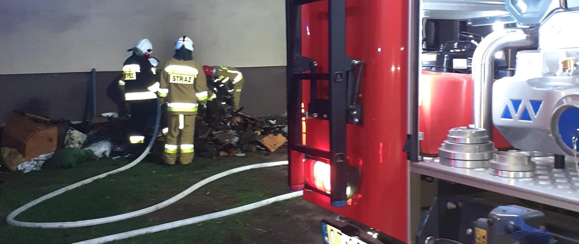 Po prawej stronie samochód pożarniczy. Po lewej strażacy pracują przy usuwaniu nadpalonego opału