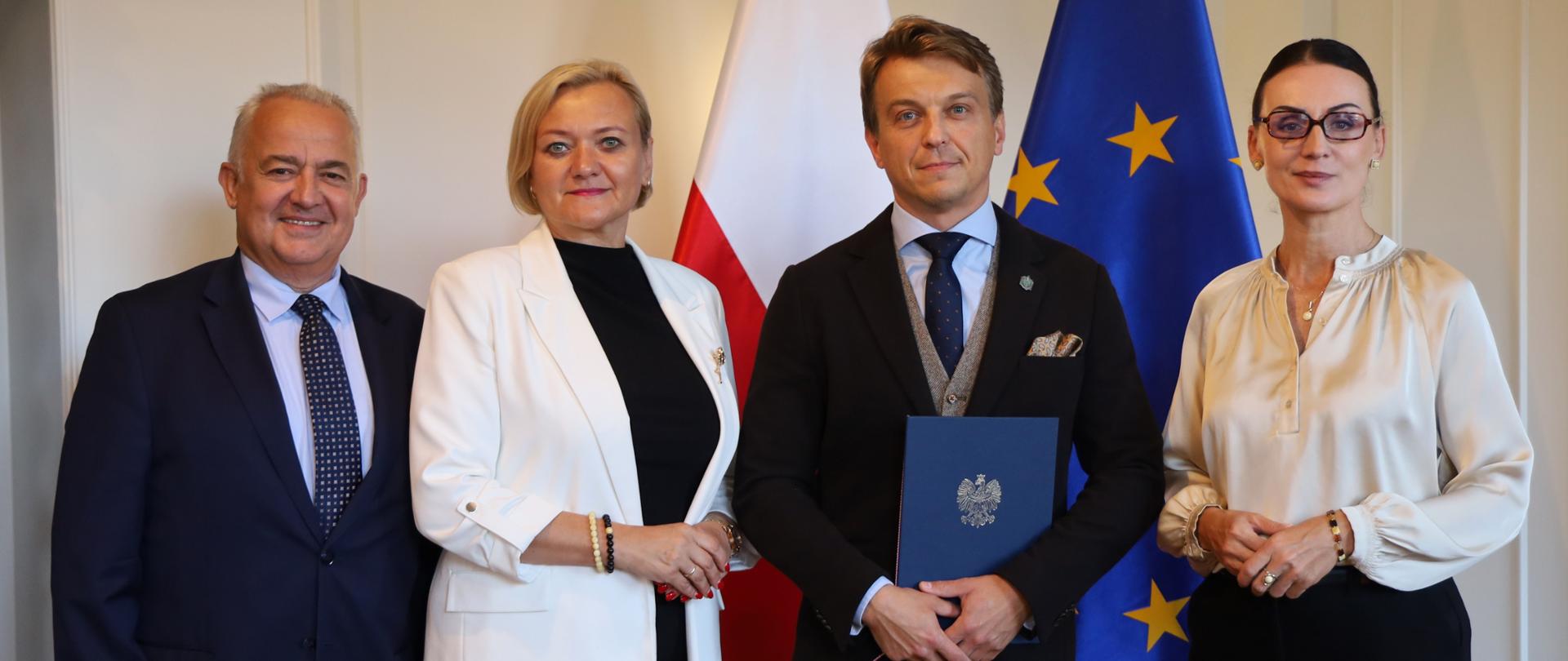 Cztery osoby pozują do zdjęcia w eleganckim wnętrzu, stojąc przed flagami Polski i Unii Europejskiej. Mężczyzna pośrodku trzyma granatową teczkę z godłem Polski.