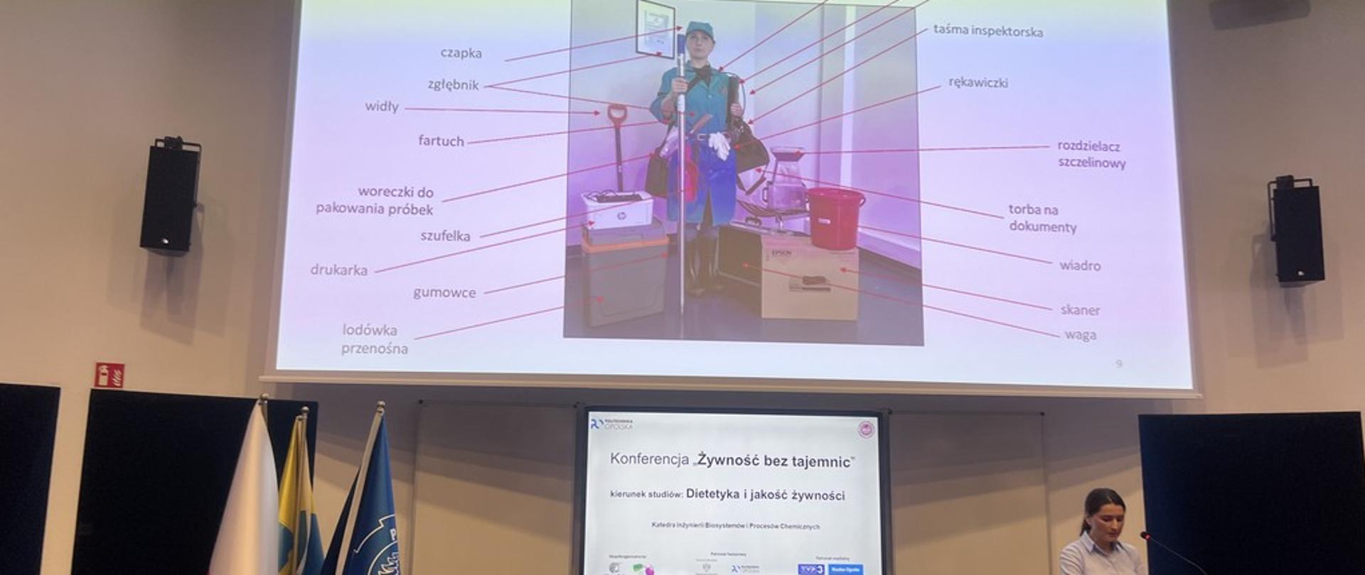 Wystąpienie przedstawicielki WIJHARS w Opolu. Nad głową prelegentki duży slajd przedstawiający inspektora WIJHARS z pełnym wyposażeniem kontrolnym, t ym zielony ubiór, sprzęt do pobierania i zabezpieczania próbek, laptop, drukarka, skaner.