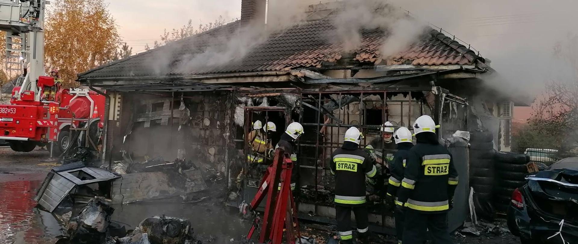 Spalony, parterowy, murowany budynek, z którego wydostaje się dym. Przed budynkiem pracują strażacy, którzy wynoszą z obiektu spalone wyposażenie. Obok budynku widoczny podnośnik samochodowy z koszem ratowniczym. 