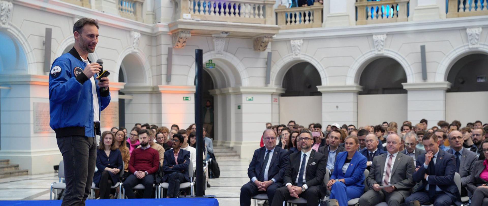 Widok z boku na salę, dr Uznański-Wiśniewski w niebieskiej kurtce stoi i mówi do trzymanego w ręku mikrofonu, za nim w rzędach krzeseł siedzą ludzie, w pierwszym rzędzie minister Kulasek.