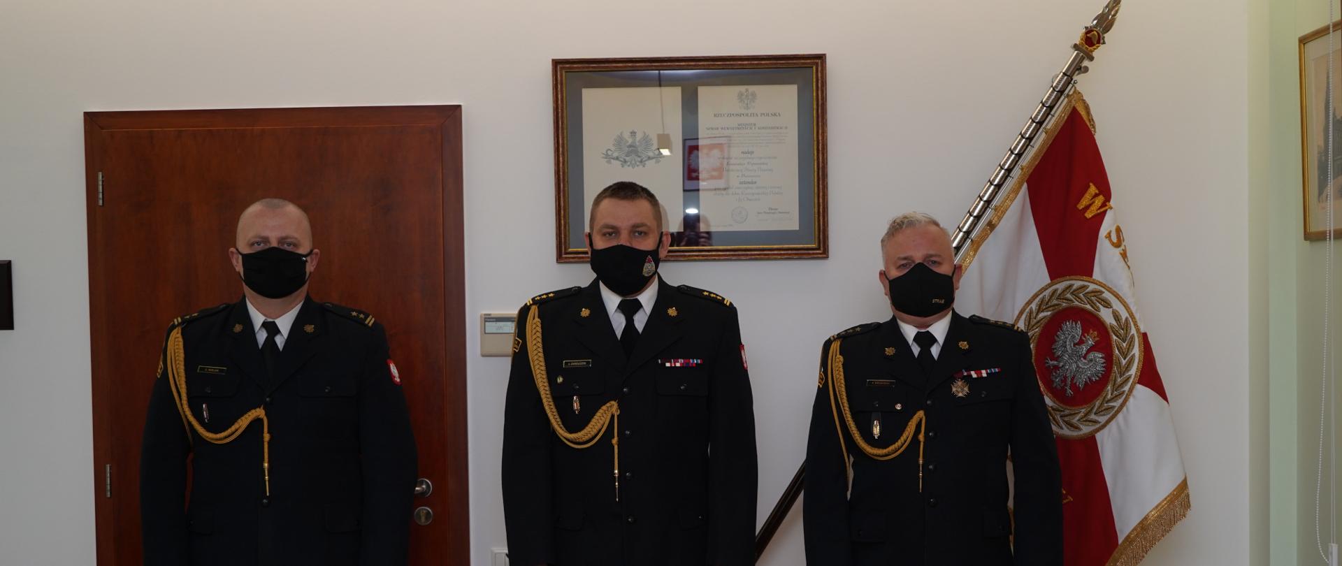 trzech strażaków stoi na baczność w ciemnych mundurach ze sznurem, białej koszuli z carnym krawatem, na twarzy mają maseczki ochronne, w tle sztandar