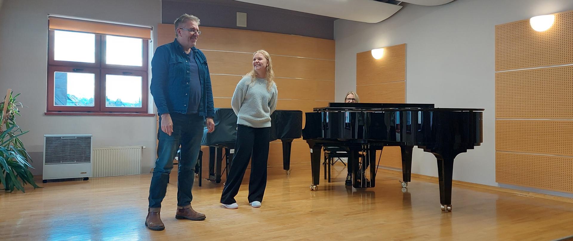 Zdjęcie na scenie sali kameralnej. Uczennica stoi na scenie wpatrzona na prowadzącego, który stoi obok niej. Oboje się uśmiechają. Za nimi po prawej stronie dwa fortepiany. Jeden przykryty pokrowcem, przy drugim pod ścianą siedzi akompaniatorka. Następnie okno przysłonięte roletą. Pod oknem kaloryfer oraz nawilżacz powietrza oraz palma w doniczce.