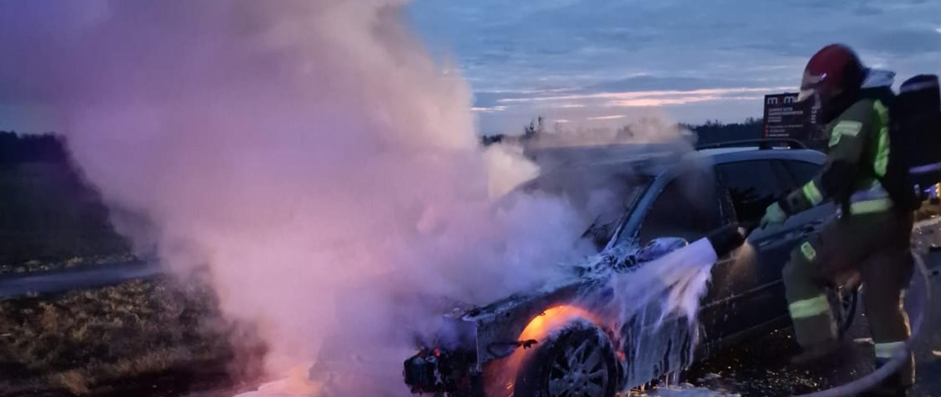 Widać dym i płomienie przy samochodzie, po prawej strażaka gasi pojazd