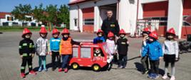 Zdjęcie przedstawia dzieci oraz strażaka stojących na placu przed jednostką przy małym samochodzie strażackim