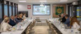 Grupa osób siedzących przy dwóch stołach konferencyjnych ogląda prezentację na ekranie w głębi sali.
