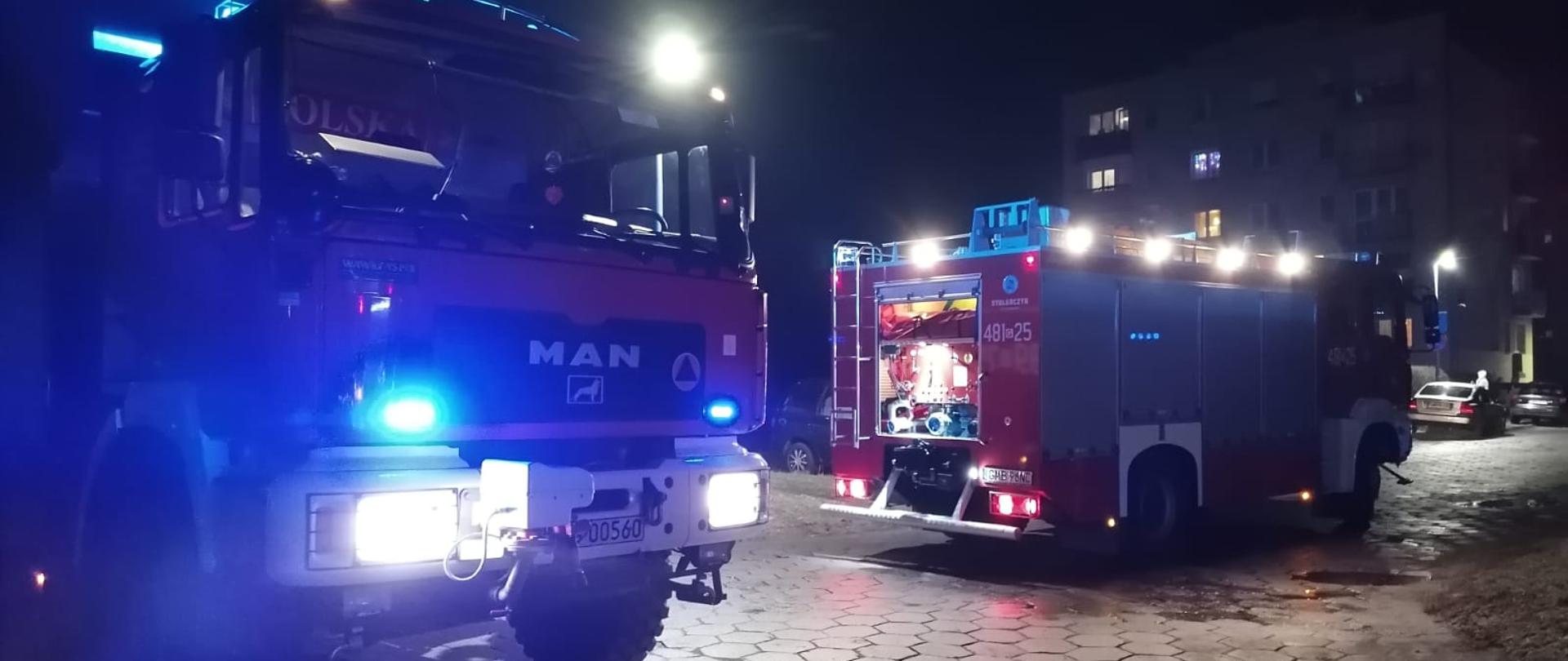 Fotografia przedstawia wozy strażackie z włączonymi sygnałami świetlnymi na jednym z osiedli w Malborku podczas akcji gaśniczej. W oddali widać blok mieszkalny na tle nocnego nieba.