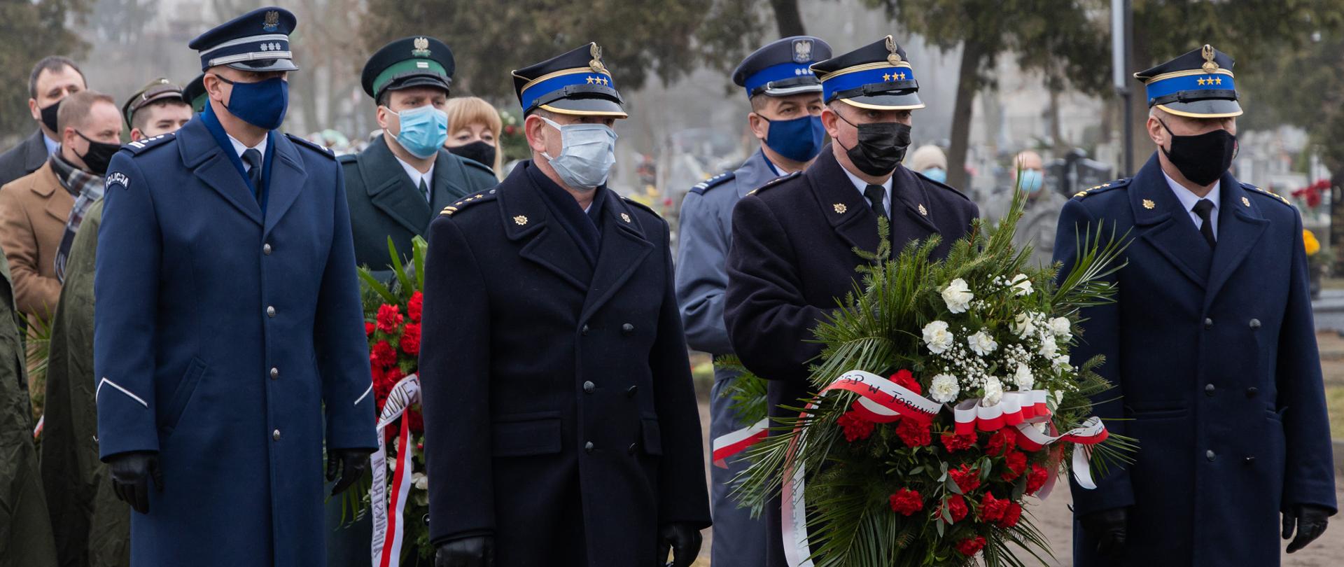 Na zdjęciu trójkami stoją delegacje służb i instytucji z wiązankami. Jako pierwsi stoją strażacy w umundurowaniu wyjściowym. Strażak w środku trzyma wiązankę, za nimi stoi delegacja policji i innych służb.