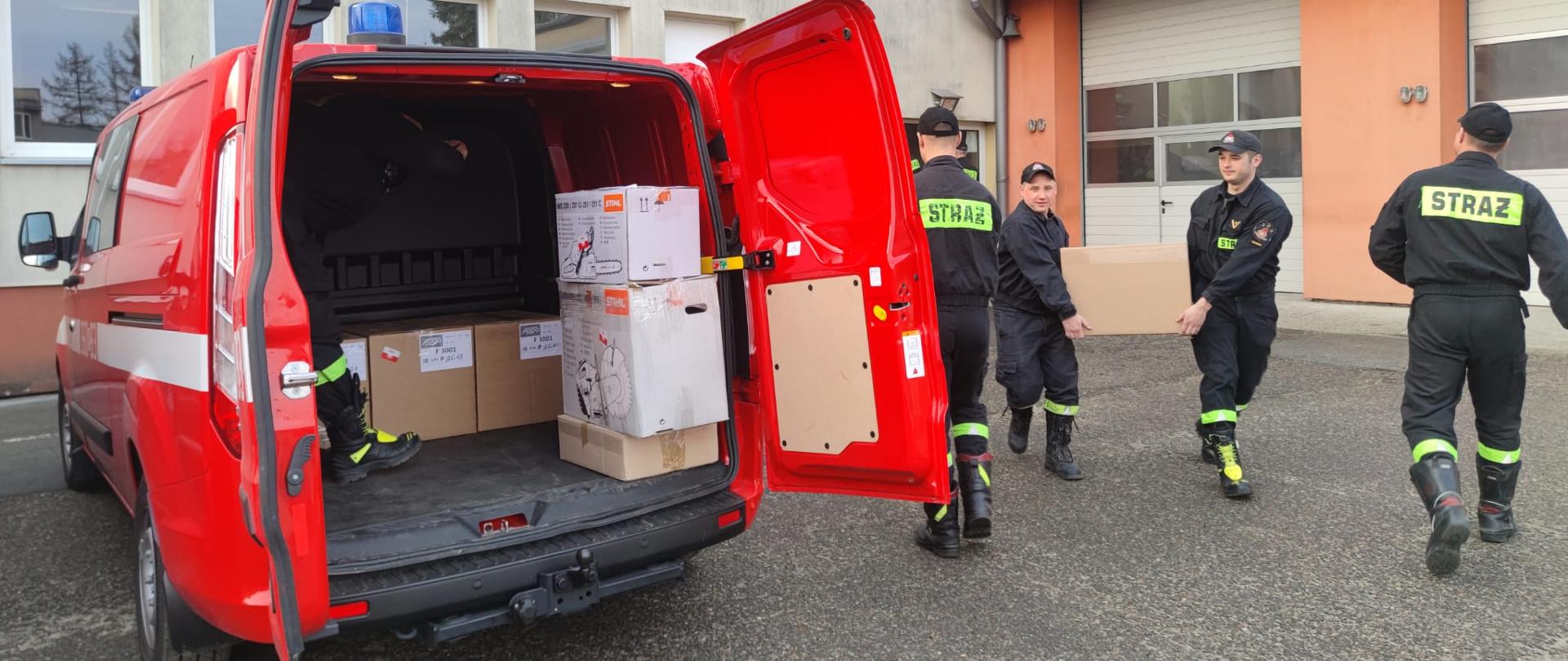 Na zdjęciu widzimy 4 strażaków podczas pakowania sprzętu do samochodu pożarniczego. Strażacy niosą kartonowe pudełko w kierunku pojazdu pożarniczego. Załadunek odbywa się na placu wewnętrznym Komendy Miejskiej Państwowej Straży Pożarnej w Tarnowie.