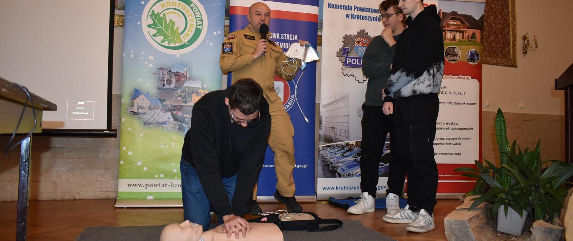 Zdjęcie przedstawia funkcjonariusza PSP wraz z uczniami szkoły średniej, którzy ćwiczą pierwszą pomoc przy użyciu fantoma oraz defibrylatora AED.