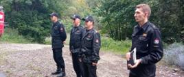 Na zdjęciu widoczni czterej strażacy w czarnych ubraniach dowódczo-sztabowych podczas odprawy po ćwiczeniach. Za strażakami las. 