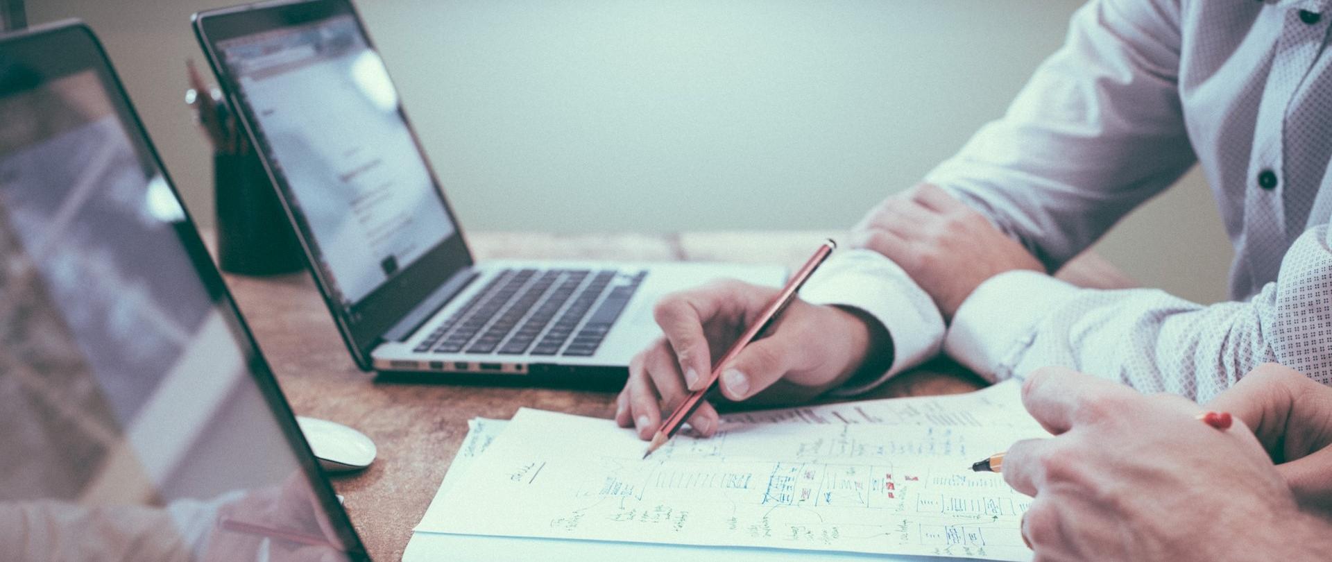 Kartki z notatkami leżące miedzy laptopami, na której dwie postacie wykazują zapisy.