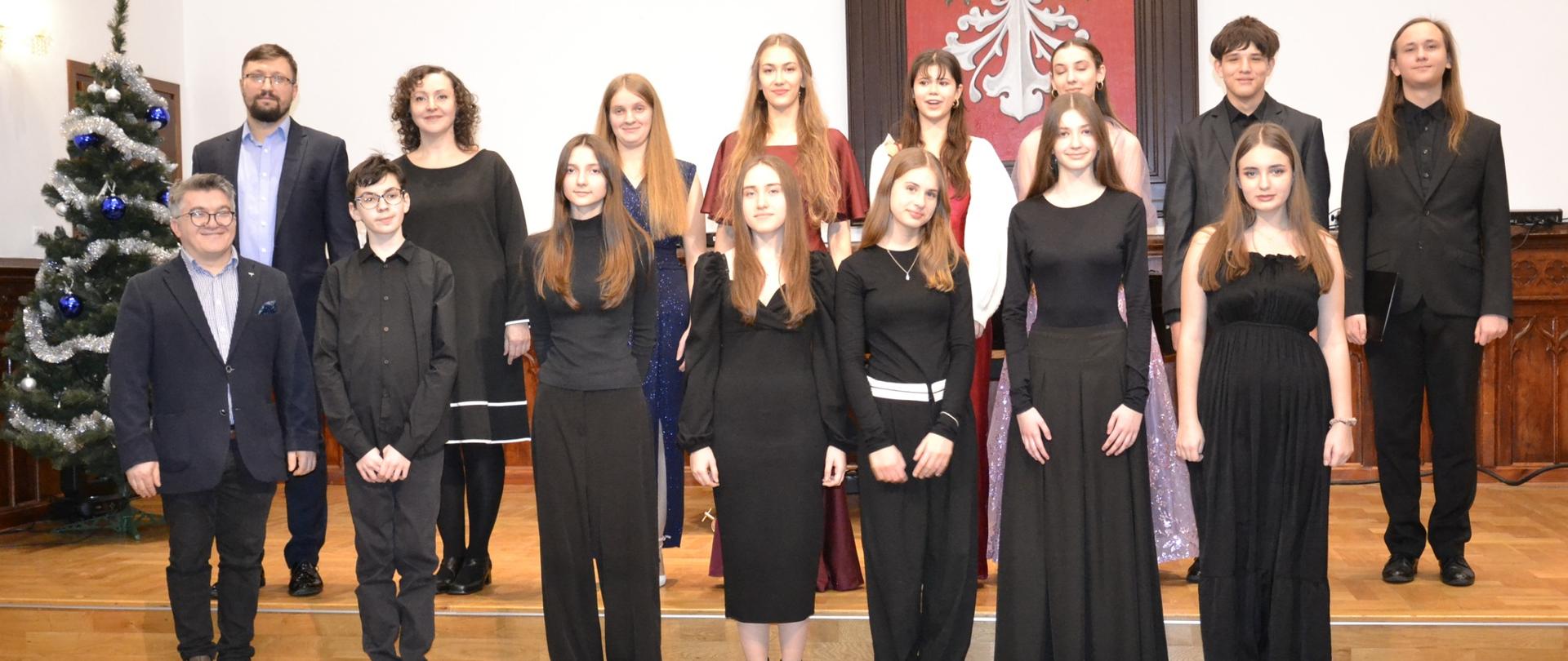 Grupa uczniów klas wokalnych stoi na scenie Sali Królewskiej po koncercie kolęd, pozując do wspólnego zdjęcia z nauczycielami.