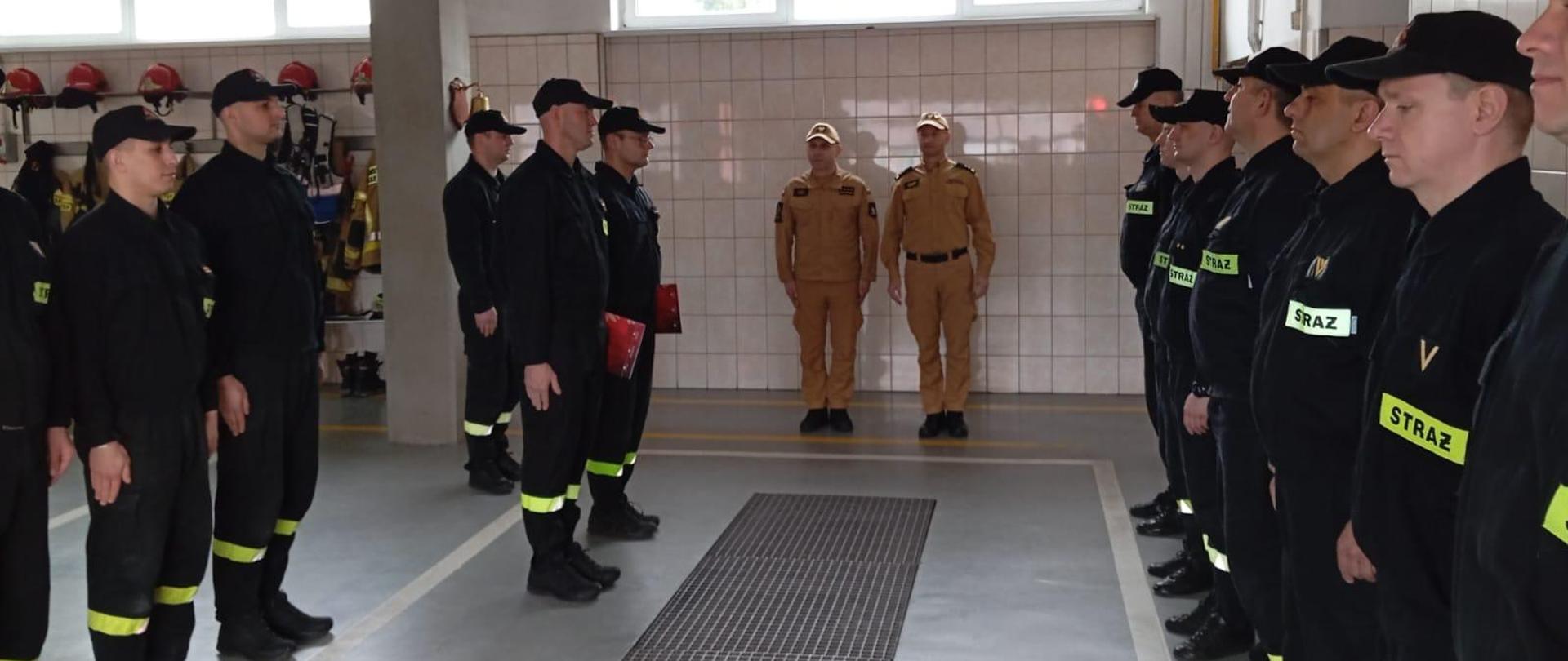 W garażu, w dwuszeregu stoją strażacy ubrani w mundury koszarowa, dwóch z nich w żółtych mundurach stoi obok siebie
