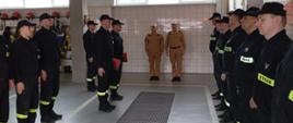 W garażu, w dwuszeregu stoją strażacy ubrani w mundury koszarowa, dwóch z nich w żółtych mundurach stoi obok siebie