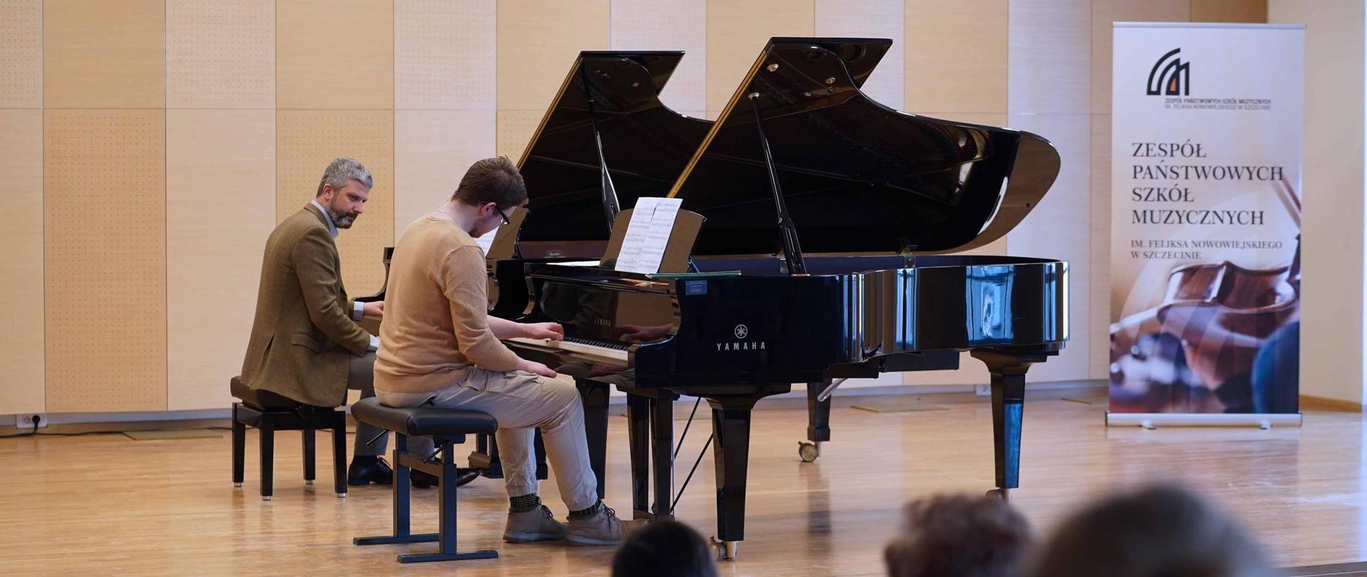 zdjęcie przedstawia ucznia wykonującego ćwiczenie w duecie razem z wykładowcą przy dwóch fortepianach na scenie sali koncertowej