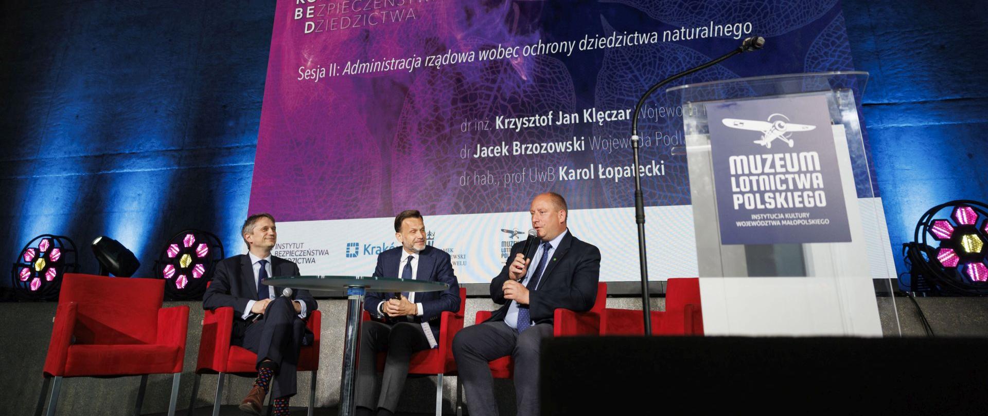 Scena, na której siedzą prelegenci, m.in. wojewoda podlaski i wojewoda małopolski. W tle duży ekran z nazwą konferencji i nazwiskami prelegentów. 