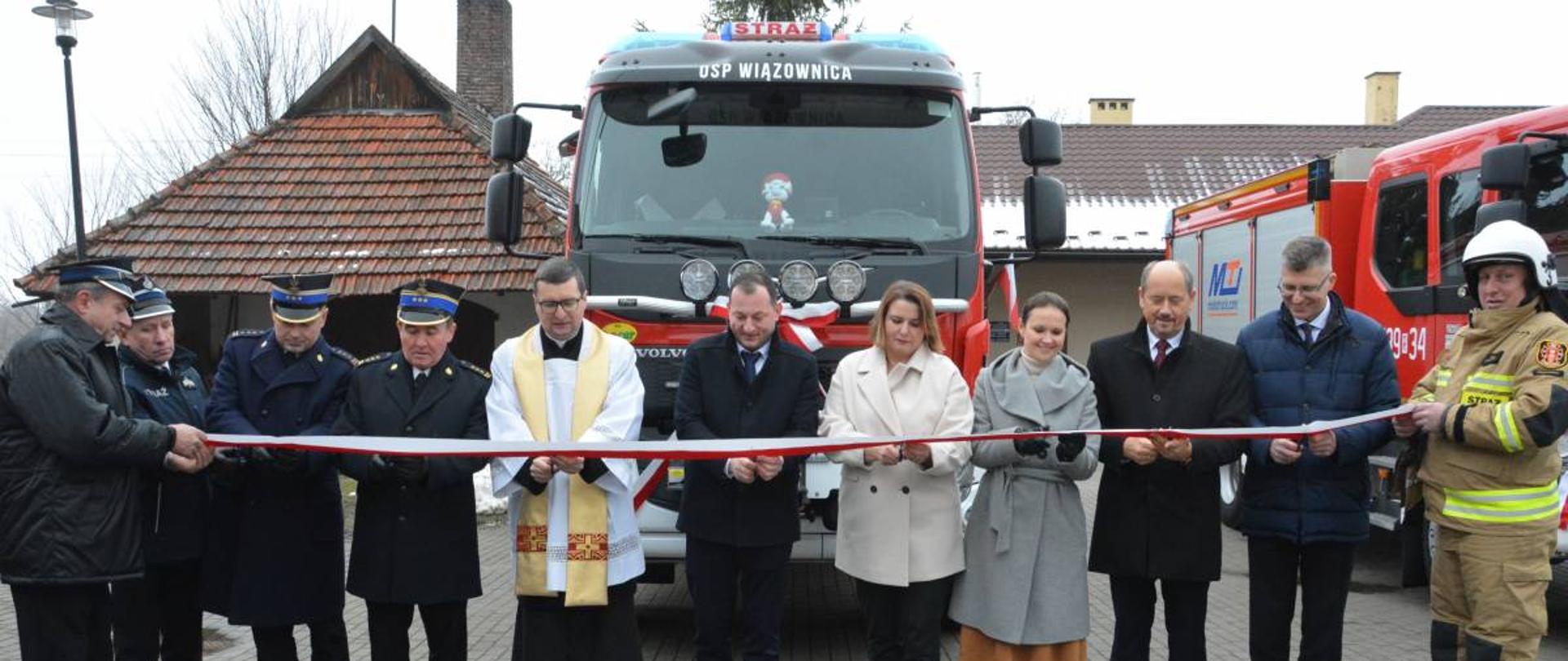 Na zdjęciu widzimy zaproszonych gości, którzy przecinają wstęgę przed nowym samochodem ratowniczo-gaśniczym. Funkcjonariusze PSP oraz druhowie OSP występują w umundurowaniu wyjściowym. W tle widoczny jest budynek.