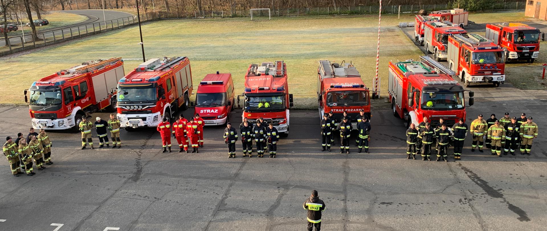 Zdjęcia z lotu ptaka w dół na plac na którym stoją strażacy na tle samochodów strażackich na dalszym planie trawnik i las