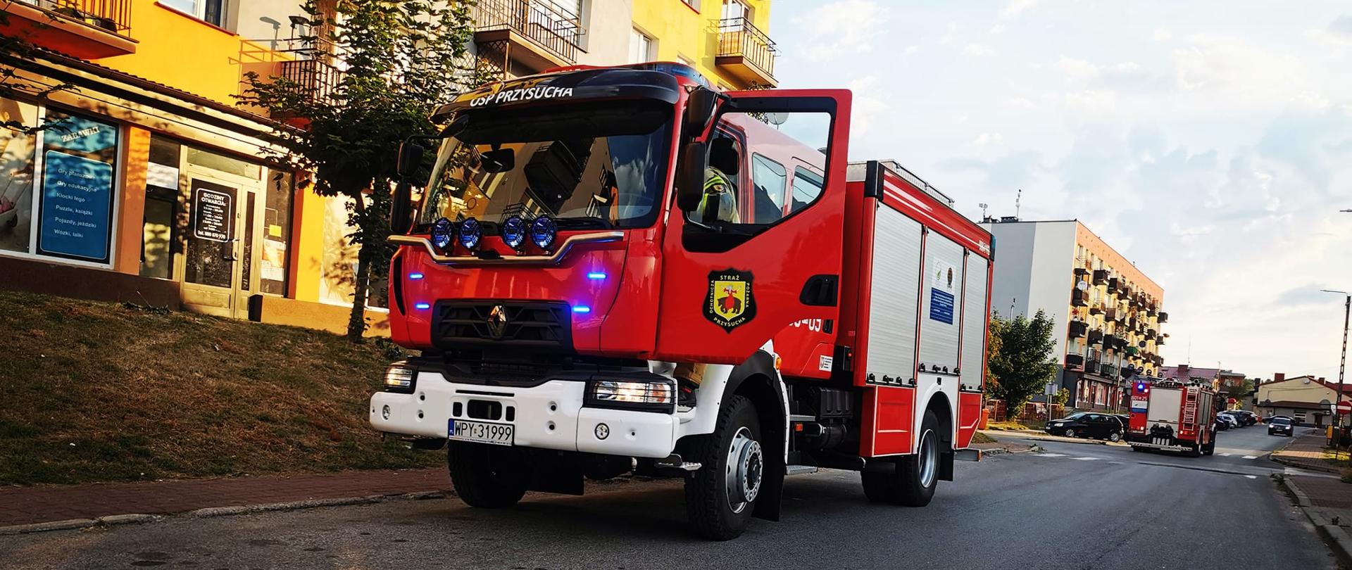 Zdjęcie przedstawia czerwony wóz strażacki z otwartymi drzwiami kierowcy, stojący na ulicy w Przysusze. Na dachu kabiny pojazdu widnieje napis "OSP PRZYSUCHA", a na drzwiach logo straży pożarnej. Włączone są niebieskie światła sygnalizacyjne. W tle widać budynki mieszkalne z balkonami, drzewa i inne samochody na drodze. Słońce świeci nisko, rzucając długie cienie i oświetlając budynki ciepłym światłem. W oddali widać inny wóz strażacki.