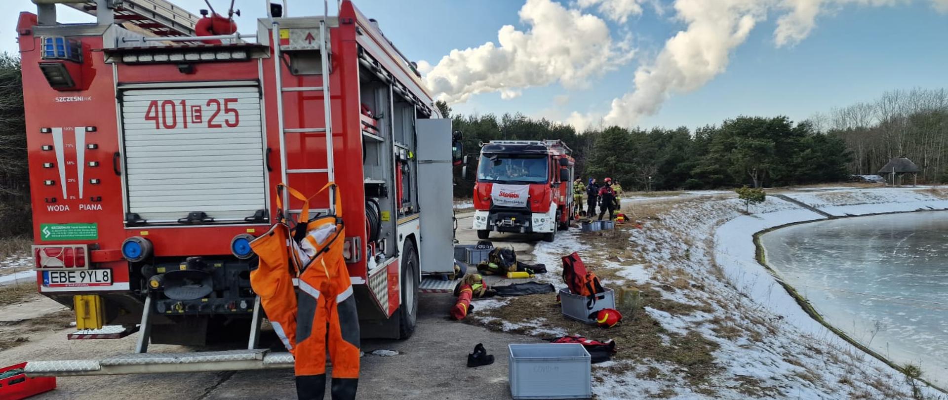 na zdjęciu widoczne samochody ciężarowe straży pożarnej wraz ze sprzętem do ratownictwa wodnego przy zamarzniętym akwenie wodnym, w oddali strażacy przygotowujący się do ćwiczeń w tle drzewa i niebo