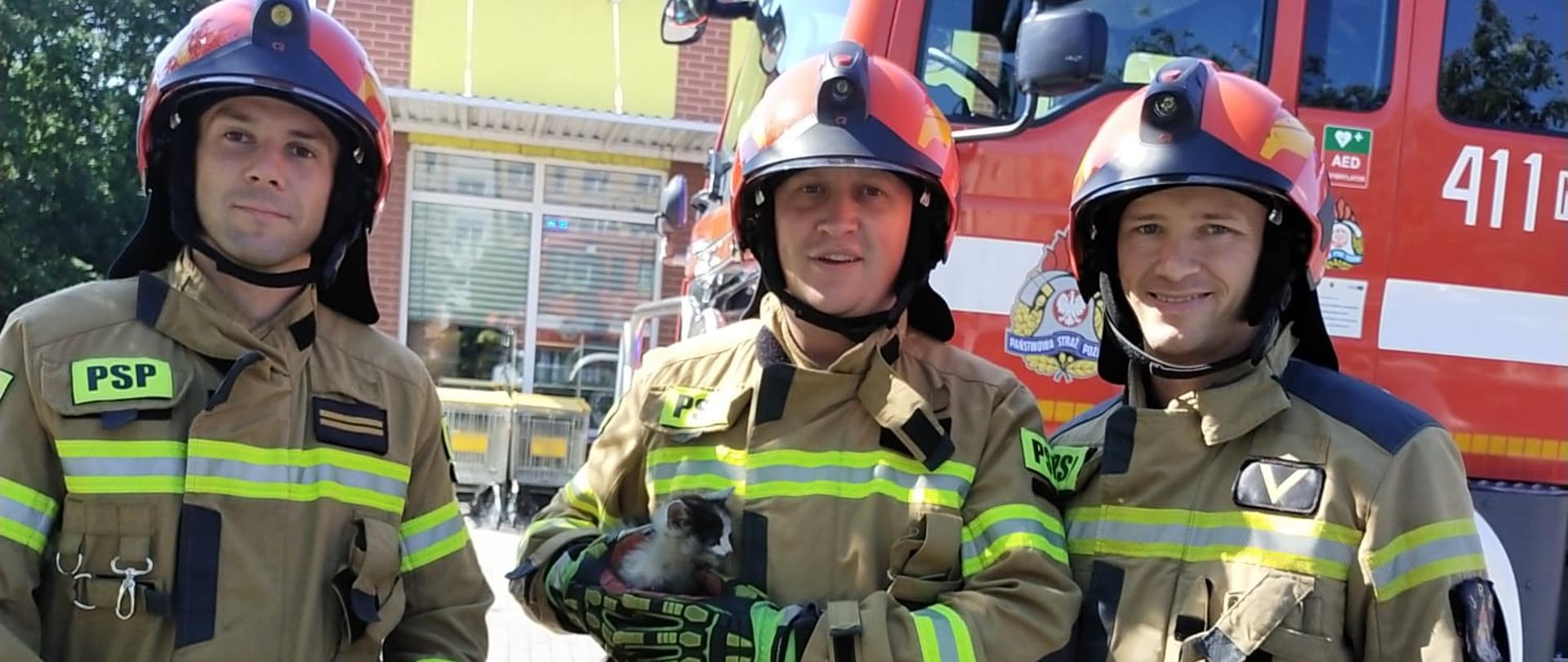 trzech strażaków w mundurach i czerwonych hełmach stoją na placu. Środkowy strażak w rekach trzyma małego kota. Z tyłu czerwony samochód strażacki.