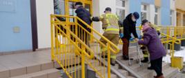 Osoby w podeszłym wieku przy pomocy strażaków wchodzą po schodach do przychodni lekarskiej. W tle kolorowa elewacja budynku.