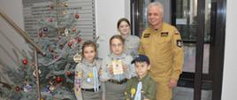 Na korytarzu komendy powiatowej PSP w Rawiczu do wspólnego zdjęcia pozują zuchy wraz z zastępcą komendanta powiatowego PSP. Wszyscy maja na sobie mundury. Chłopiec - zuch na głowie ma czapke dzokejkę. W rękach trzyma lampion, z Betlejemskim Światłem Pokoju. Dziewczynka stojąca obok ma w rękach tabliczke z życzeniami. Z lewej strony stoi ubrana w ozdoby, choinka.