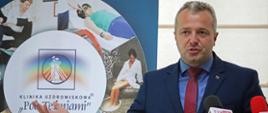 Wojewoda Kujawsko-Pomorski przemawia stojąc. W tle - ścianka reklamowa kliniki uzdrowiskowej. 