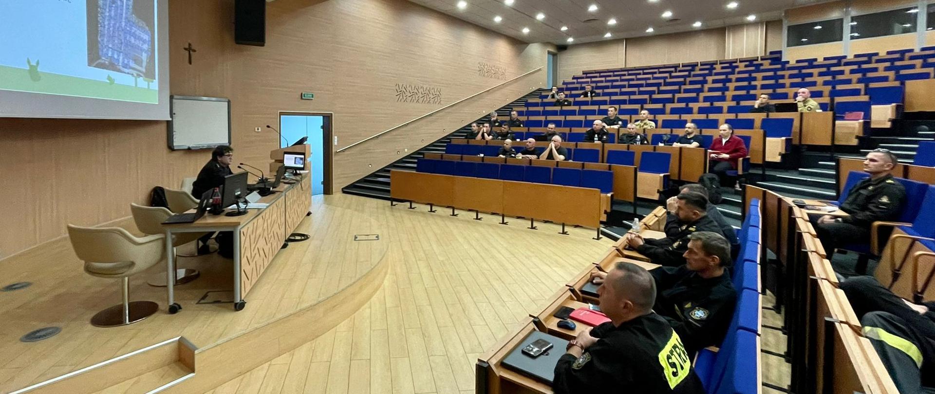 Aula wykładowa - kadr ogólny. Na pierwszym planie rzędy ławek z uczestnikami szkolenia. W głębi podwyższenie z biurkiem prowadzącego zajęcia, za którym widoczny jest ekran z prezentacją multimedialną.