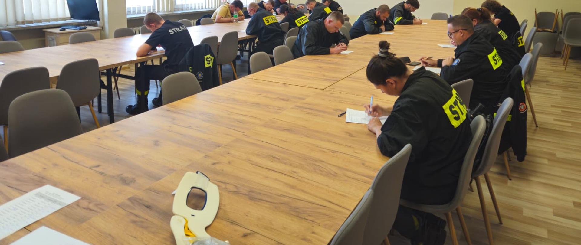 Grupa strażaków w ciemnych mundurach z napisem „STRAŻ” siedzi przy długich, drewnianych stołach w jasnej sali konferencyjnej i pisze test pisemny.