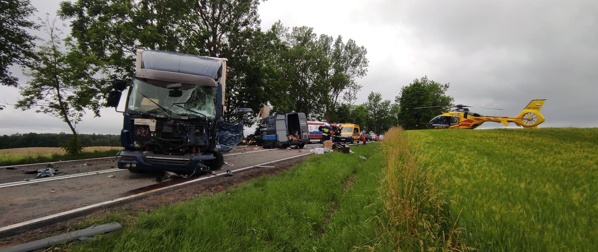 Na pierwszym planie zdjęcia po lewej stronie na jezdni znajduje się rozbity samochód ciężarowy, w oddali widać uszkodzony samochód bus oraz zespół ratownictwa medycznego. Po prawej stronie na polu widać śmigłowiec lotniczego pogotowia ratunkowego.