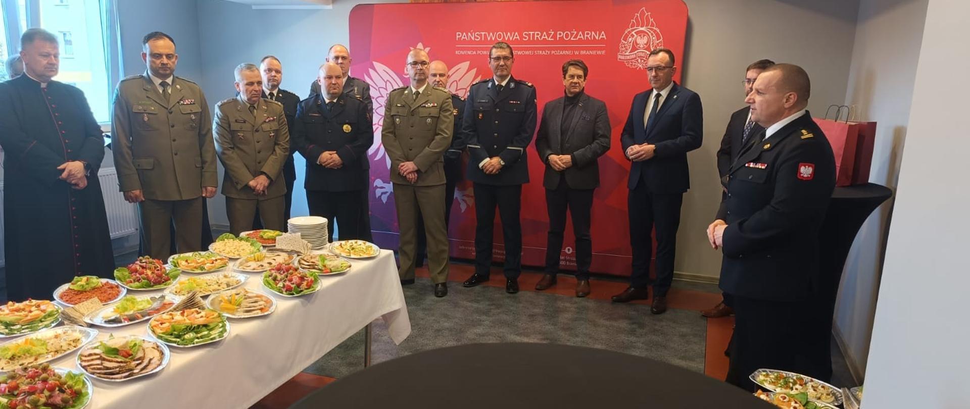 Uroczyste potkanie na świetlicy. Na zdjęciu grupa przedstawicieli służb mundurowych w mundurach wyjściowych oraz samorządowcy. Z lewej strony stoi stół z potrawami.