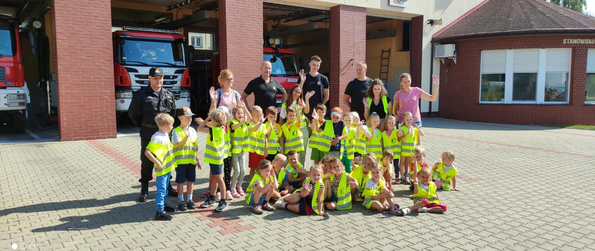 Zdjęcie przedstawiające grupę przedszkolaków wraz ze swoimi opiekunami w czasie wizyty u średzkich strażaków. W tle budynek Komendy Powiatowej PSP w Środzie Wielkopolskiej.