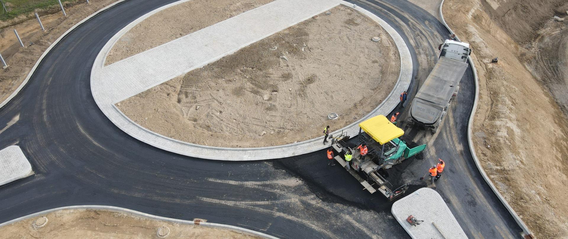 DK9 Ostrowiec Świętokrzyski - zdjęcie z drona, układanie podbudowy asfaltowej na rondzie, dwa samochody ciężarowe, rozściełacz, pracownicy w odblaskowych kamizelkach 