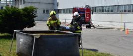 Zdjęcie przedstawia strażaków w trakcie wkładania butli z acetylenem do zbiornika ppoż. z wodą w celu schłodzenia butli na tle zakładu i pojazdu pożarniczego