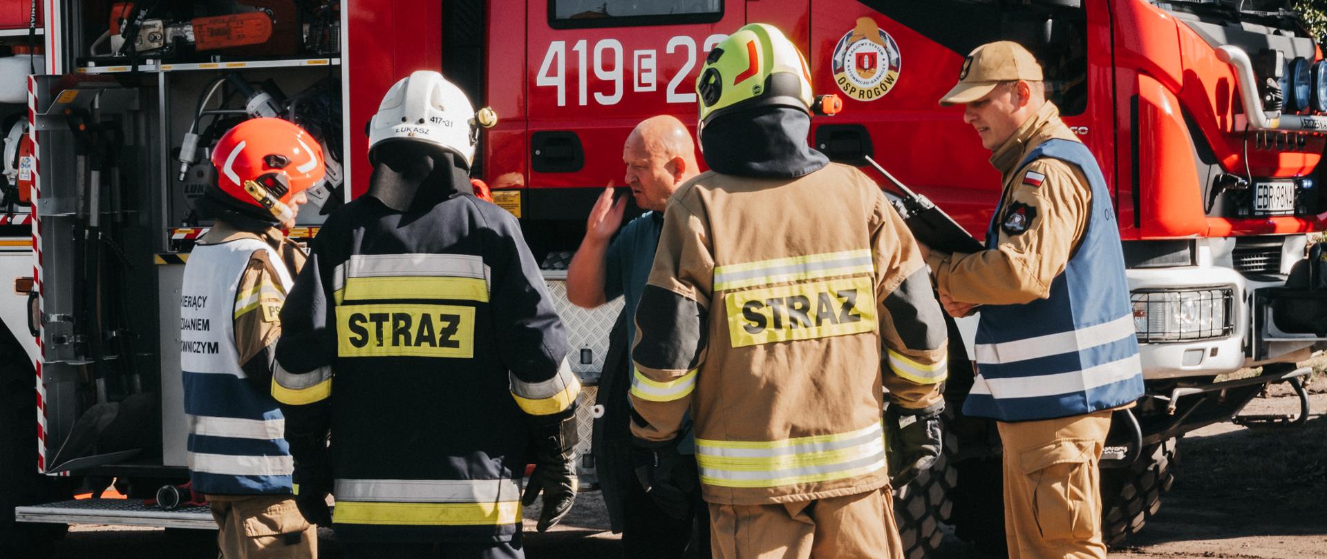 na terenie tartaku w rogowie trwa odprawa strażaków oraz kierującego działaniem ratowniczym na tle wozu strażackiego