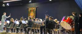 Zdjęcie przedstawia młodych muzyków z instrumentami dętymi oraz perkusją, oraz postać dyrygenta z batutą - na sali koncertowej. 