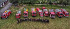 Widok z drona na samochody pożarnicze oraz strażaków ochotników zebranych na boisku w Chociczy podczas ćwiczeń z gaszenia pożaru lasu