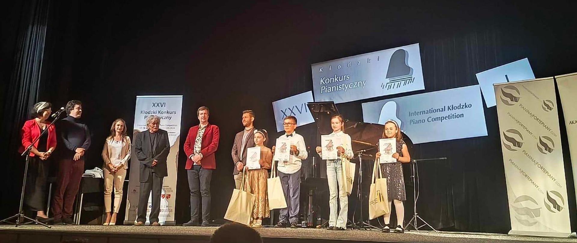 Stojący uczestnicy konkursu wraz z jurorami na scenie Kłodzkiego Ośrodka Kultury, którzy trzymają dyplomy za szczególne osiągnięcia w konkursie