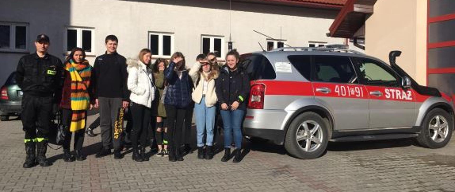 Dzieci na tle samochodu po lewej stronie strażak w umundurowaniu służbowym, w oddali budynek komendy 